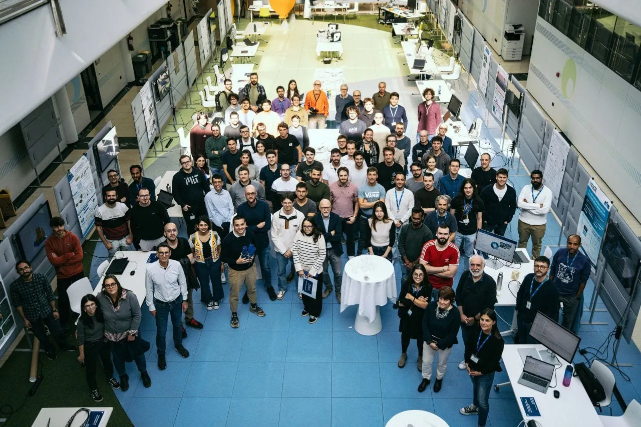 A large group of people photographed from above in a long exhibition hall with demo tables.