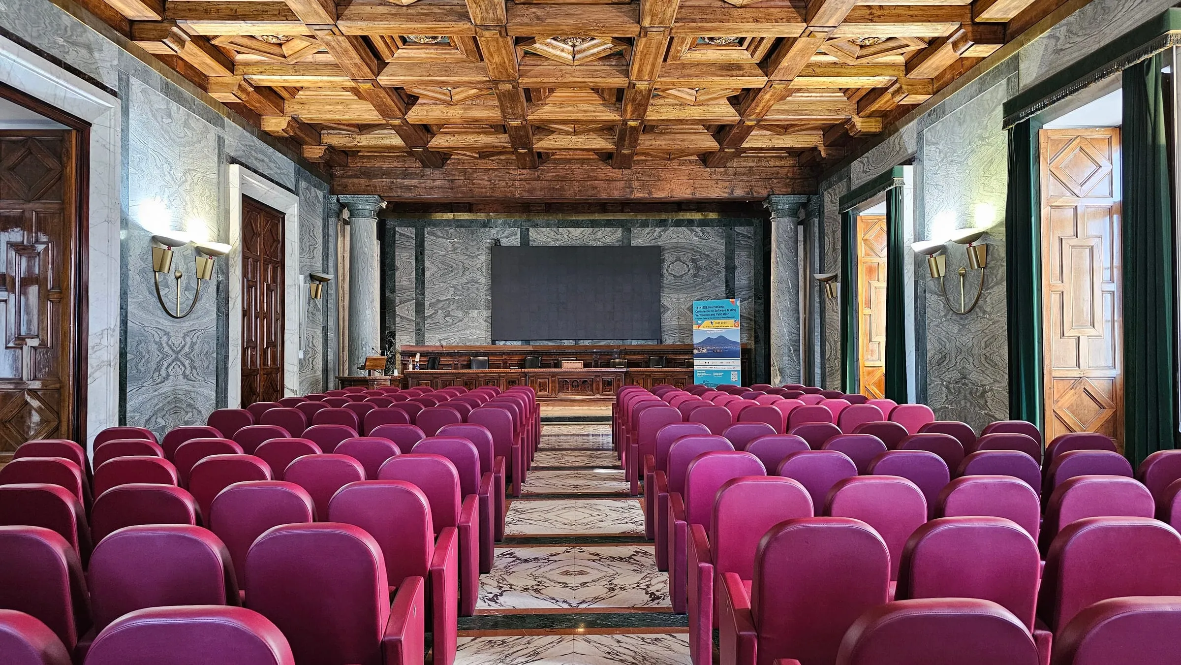 Rows of red velvet seats facing the stage in the conference hall, under an ornate wooden ceiling.