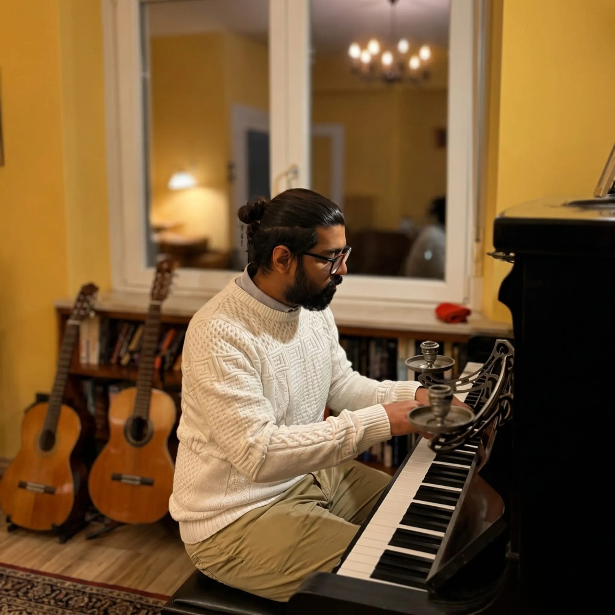 Khandaker Rahin playing an upright piano at home in a cream sweater, two acoustic guitars against the wall behind.