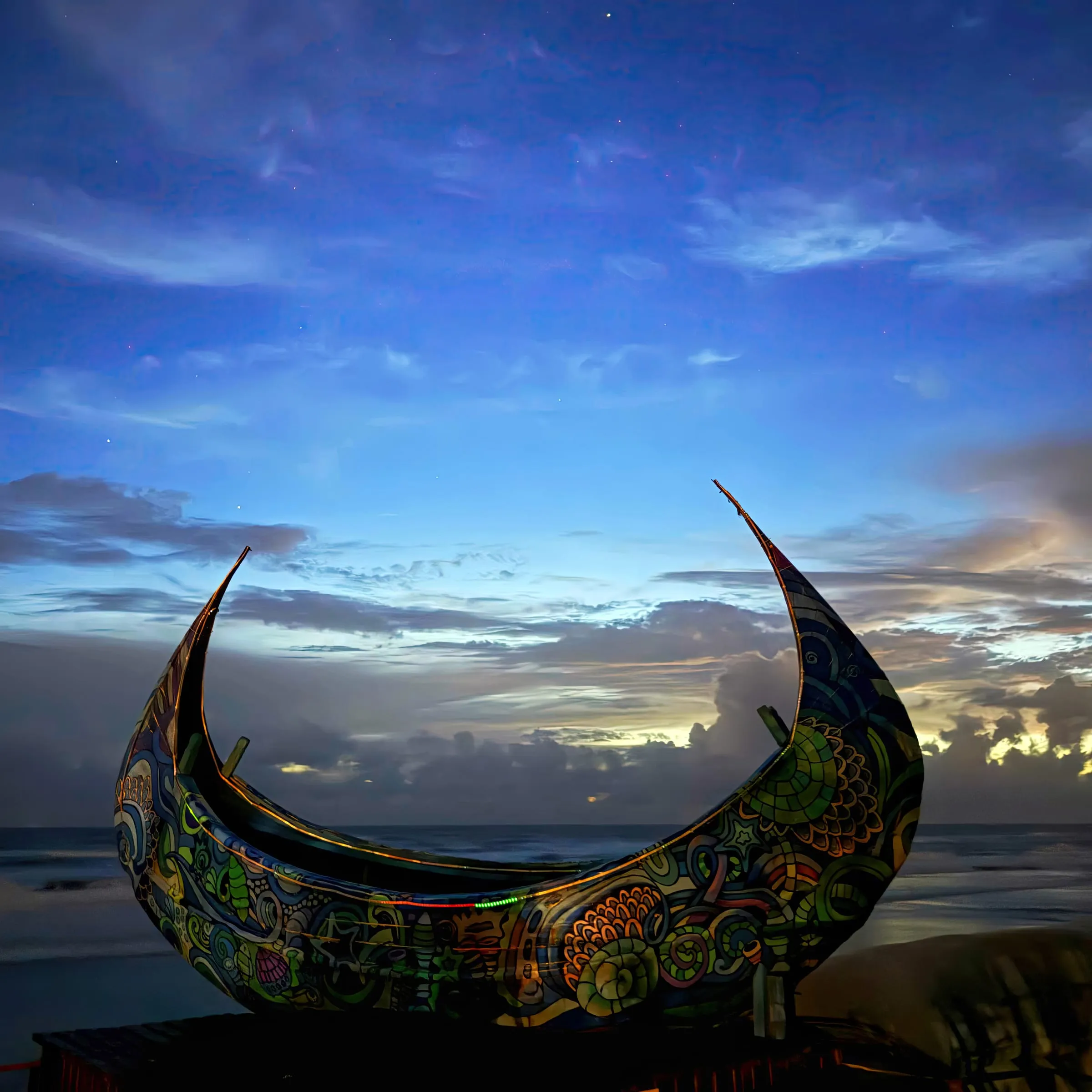 Wind of Bliss artwork, a sculpture shaped like a crescent boat, painted in intricate patterns, against a blue dusk sky.